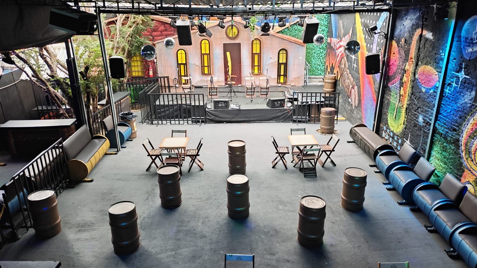 Vista completa da Villa do Som com palco cenográfico de casa de pedras, lounges de barril e mural grafitado